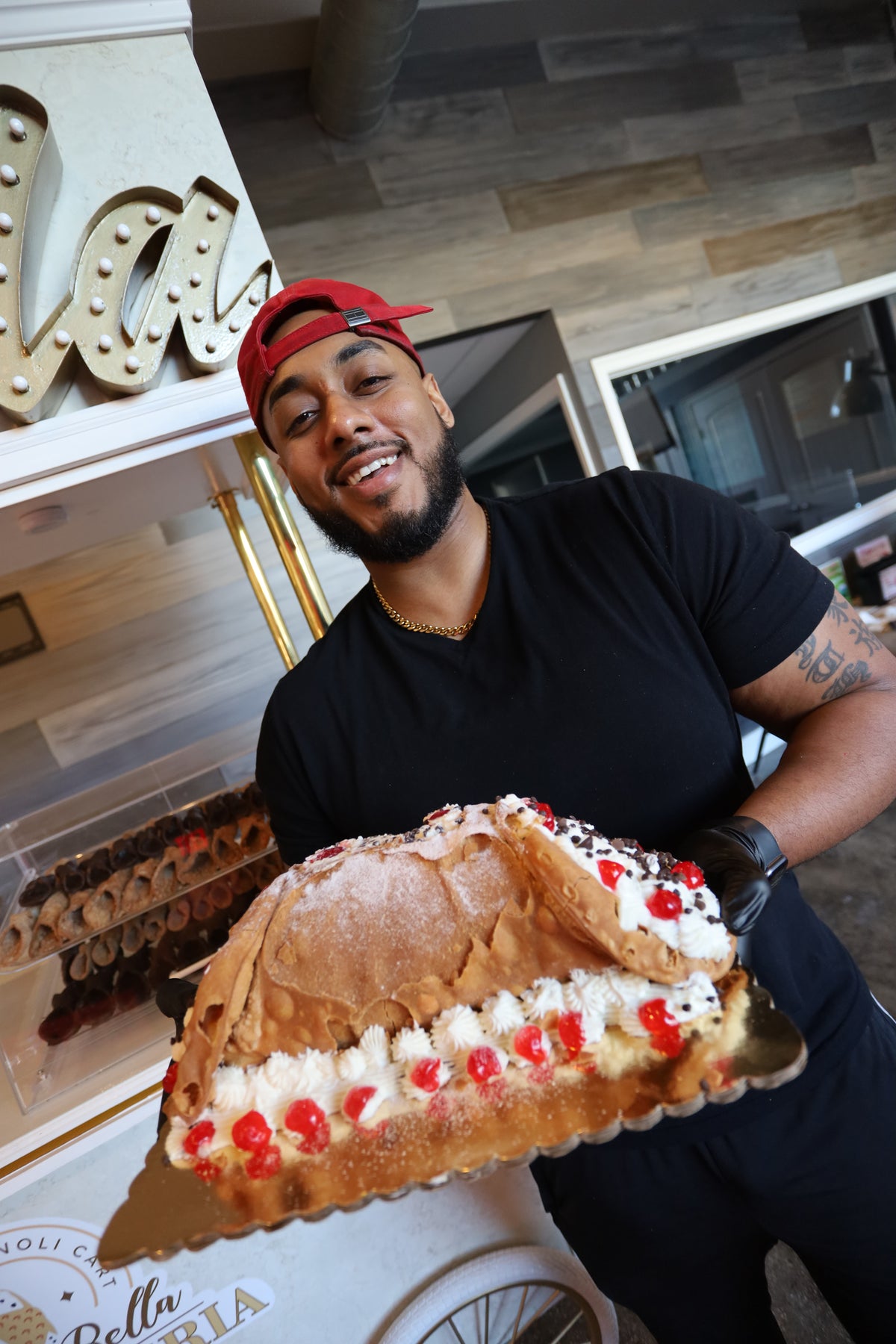 Person holding a large cannoli with red cherries and white cream in an indoor setting