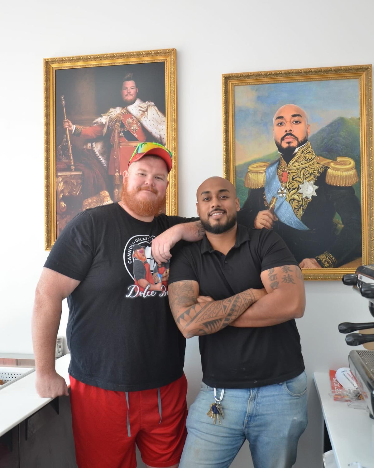 Two men standing together with portraits of historical figures on a wall behind them.