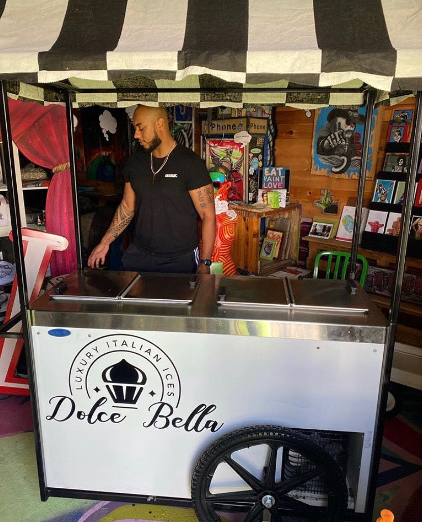 Man standing behind a Dolce Bella ice cream cart in an indoor setting with books and decor.