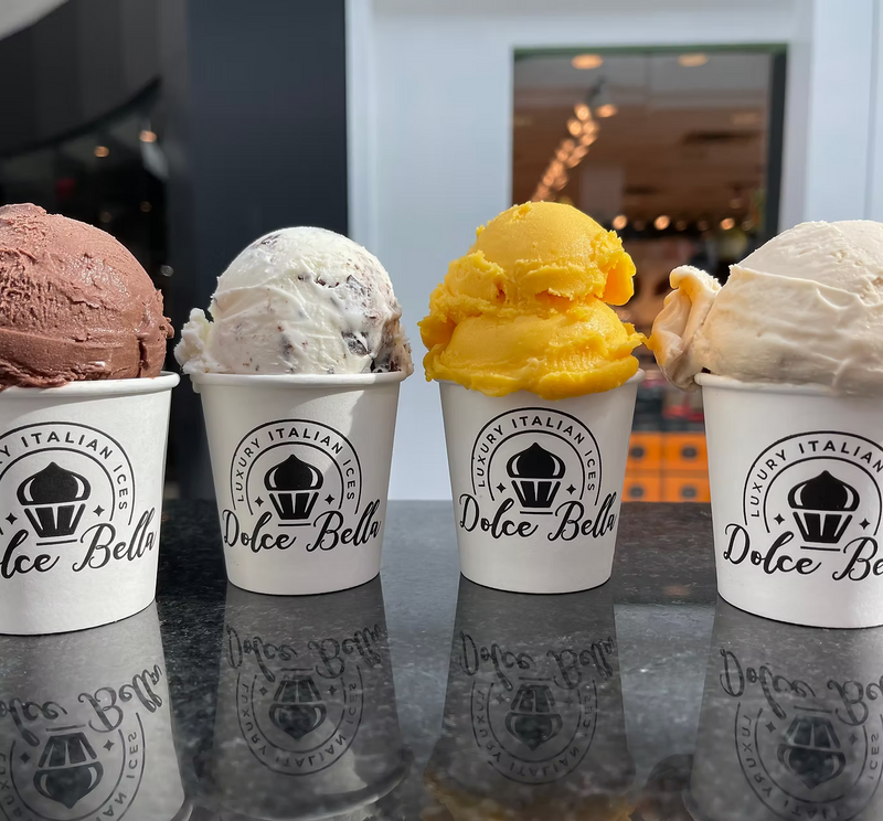 Four cups of gelato from Dolce Bella with different flavors on a reflective surface.
