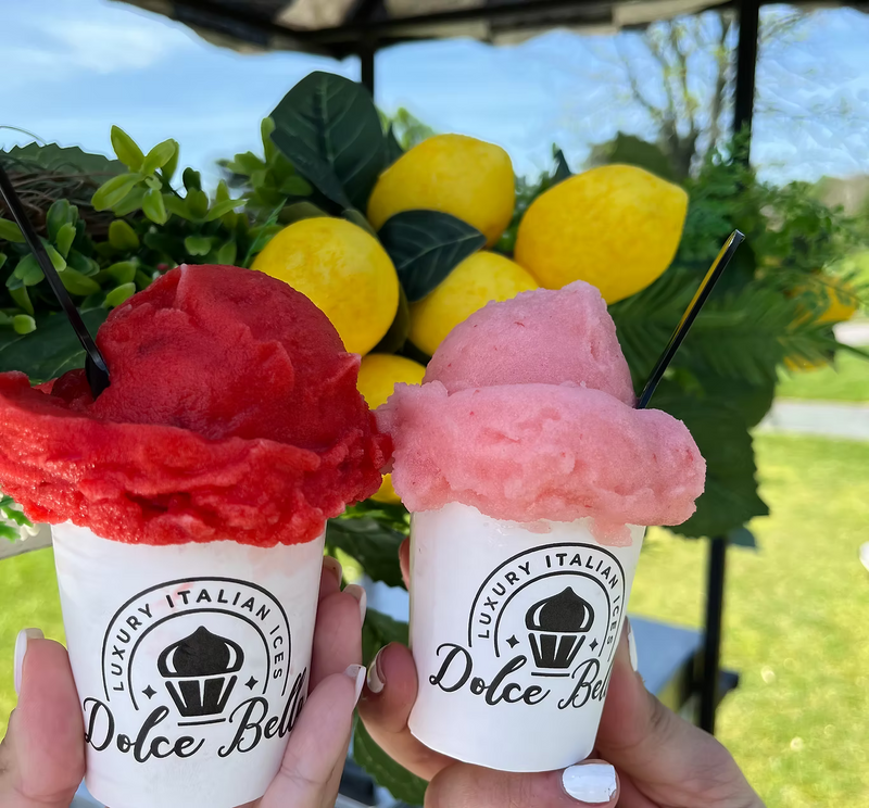 Two cups of red and pink ice cream with 'Dolce Bella' branding, held in front of lemons and greenery.